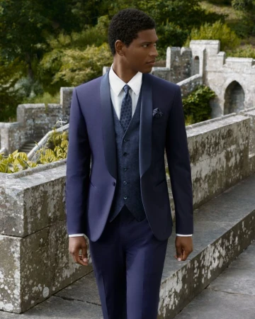 Dark-skinned male model in a blue suit with vest and tie walks along a stone wall with blurred green foliage in the background.