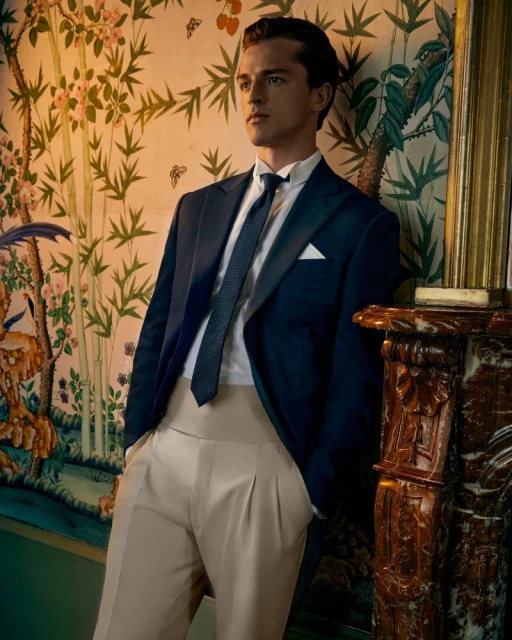 Portrait of a fair-skinned adult male wearing a navy suit and khaki pants, leaning against a marble fireplace indoors, floral wallpaper.
