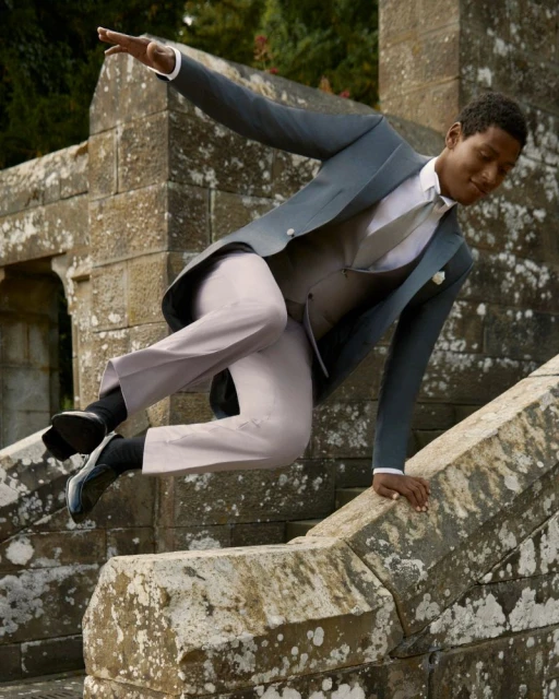 Man in gray suit jumping over stone wall. Formal wear fashion shoot in outdoor setting. Male fashion model leaping in mid-air.
