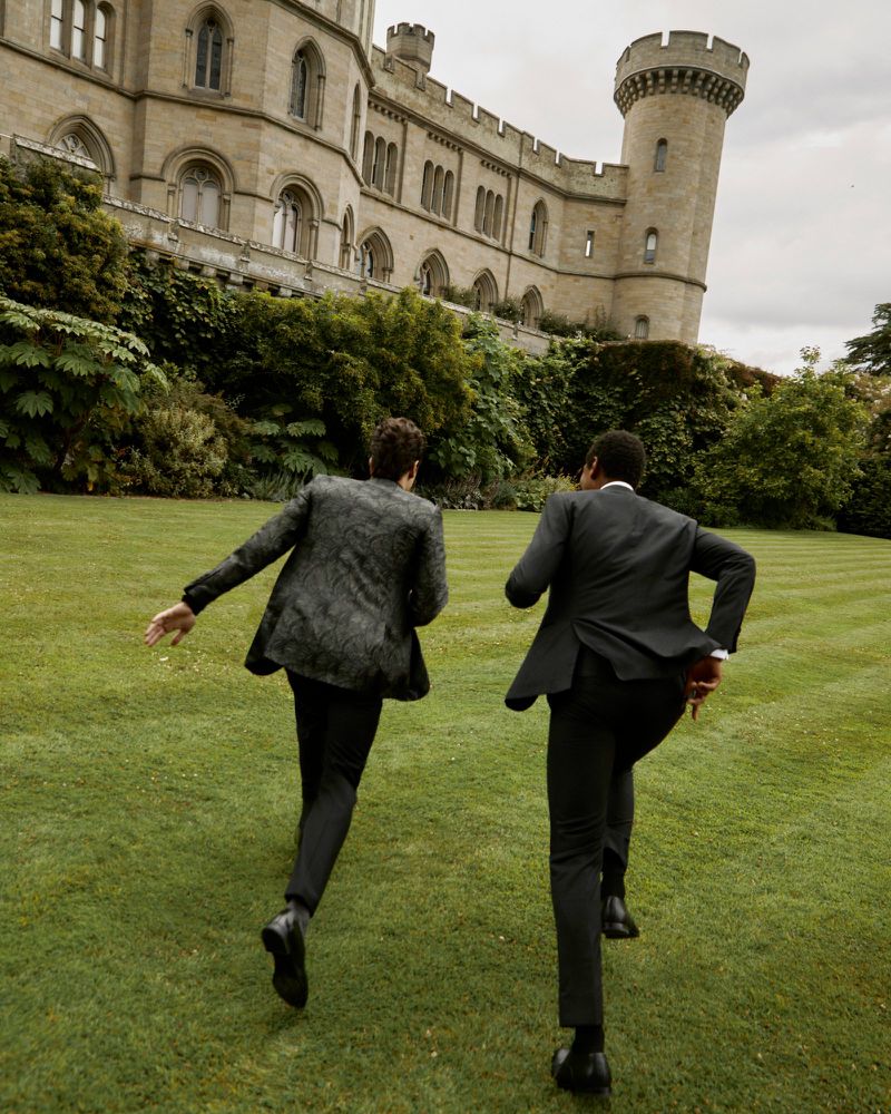 Two men in suits running on the lawn toward a castle. The suits are dark, and the lawn is green. The castle is made of stone.