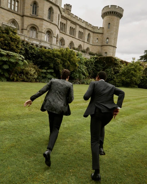 Two men in suits running on the lawn toward a castle. The suits are dark, and the lawn is green. The castle is made of stone.
