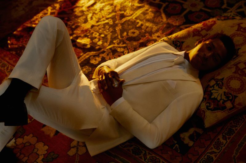 An adult Black male lies reclining on a patterned rug wearing an off-white suit and black shoes in a dimly lit indoor environment.