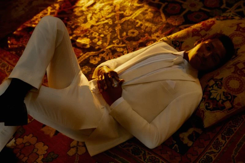 An adult Black male lies reclining on a patterned rug wearing an off-white suit and black shoes in a dimly lit indoor environment.