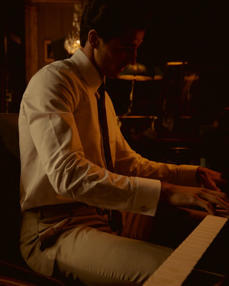 Man in suit playing piano in dim, warm light. Cream shirt, dark tie, light pants, playing piano. Warm, inviting atmosphere. Music theme.