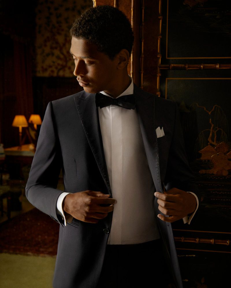 Man in a navy tuxedo with bow tie standing indoors against dark wood paneling, looking to the side with a soft, warm light.