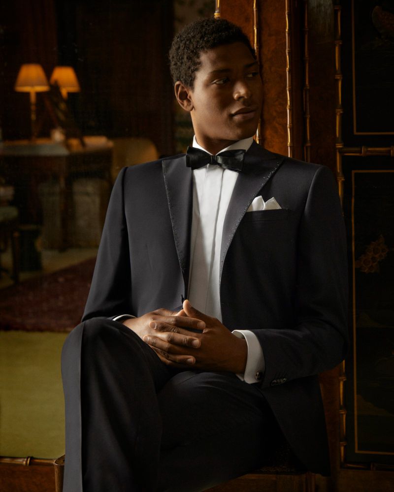 Portrait of a Black man in a classic black tuxedo with a bow tie, seated in an interior setting with soft lighting and elegant decor.