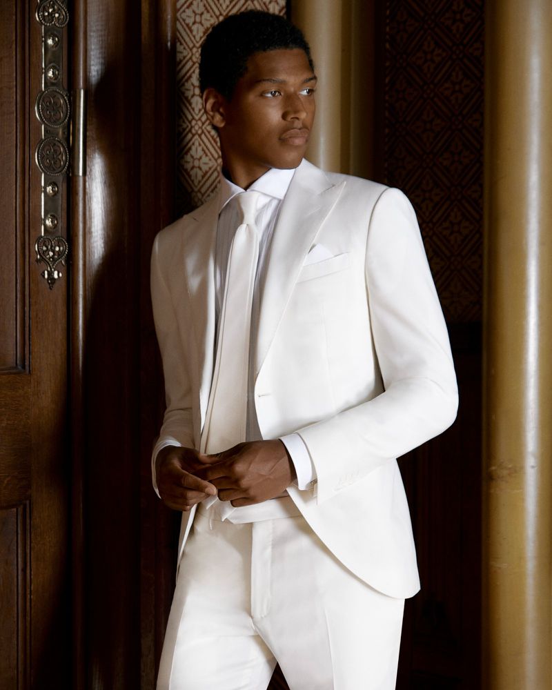 Portrait of an adult male in a white suit and tie standing indoors, featuring a neutral expression and elegant formal wear.