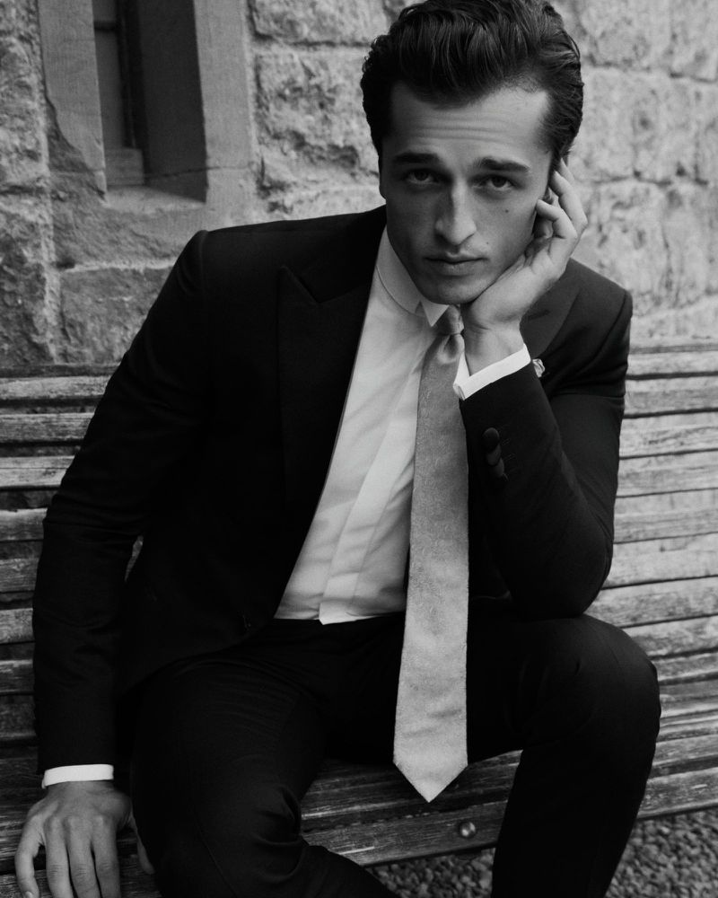 Man seated on a wooden bench wearing a dark suit jacket, white shirt, and tie with hand to his cheek