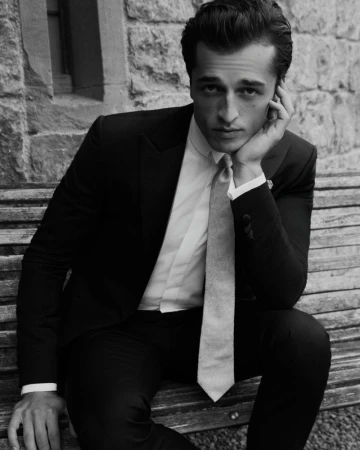 Man seated on a wooden bench wearing a dark suit jacket, white shirt, and tie with hand to his cheek