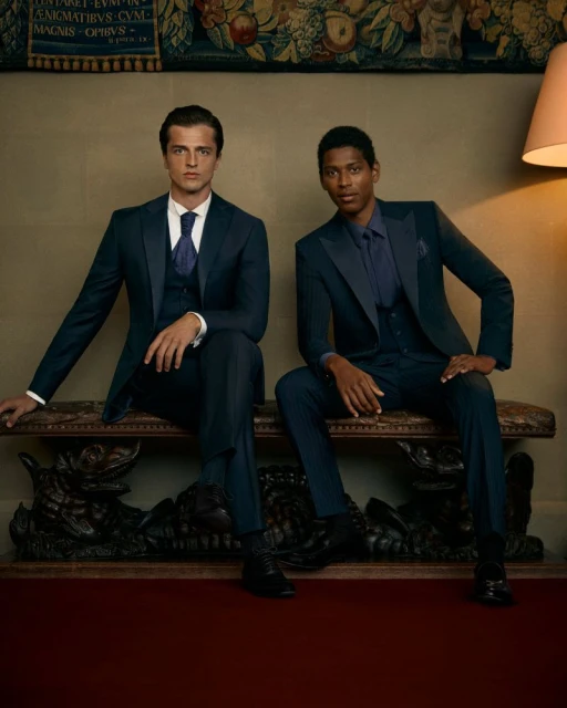 Two men in elegant blue suits pose indoors on an ornate bench against a tapestry, under warm lighting from a nearby lamp.