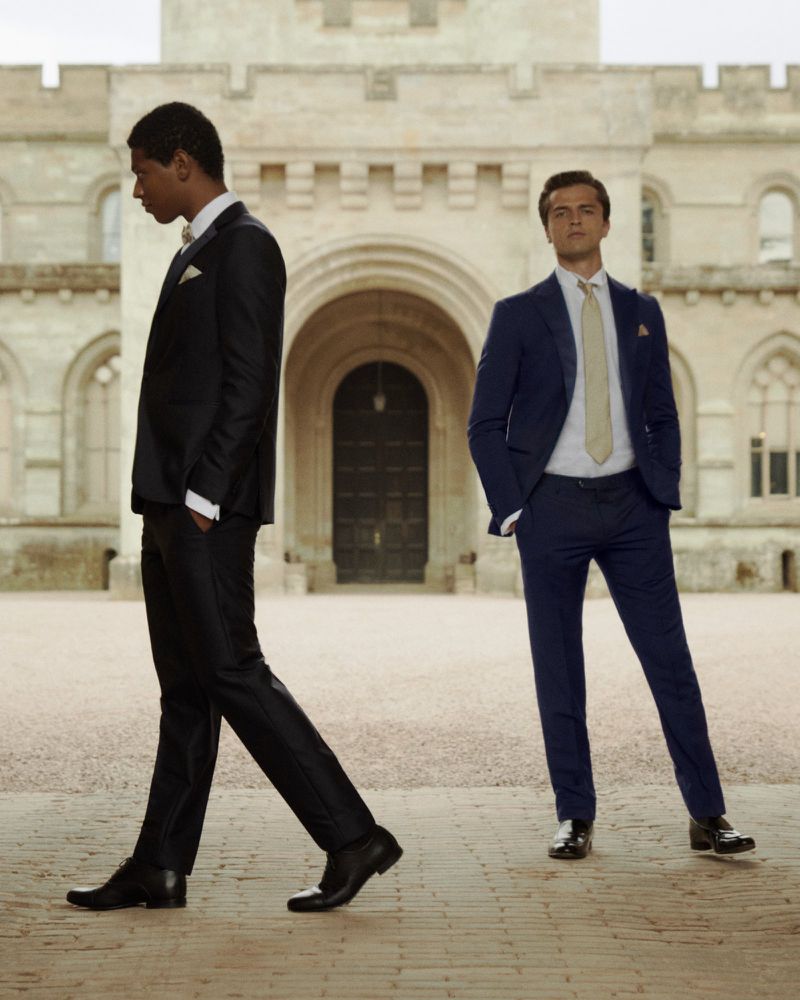 Two men in formal suits stand on a stone walkway in front of a castle, the Black man walking towards the left and the Caucasian man standing.