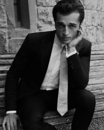 Man seated on a wooden bench wearing a dark suit jacket, white shirt, and tie with hand to his cheek