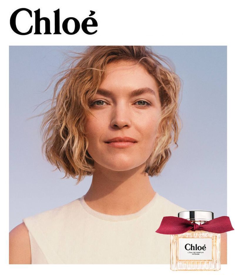 Beauty shot of a blonde woman with short hair and a perfume bottle with red ribbon, set against a blue gradient background.