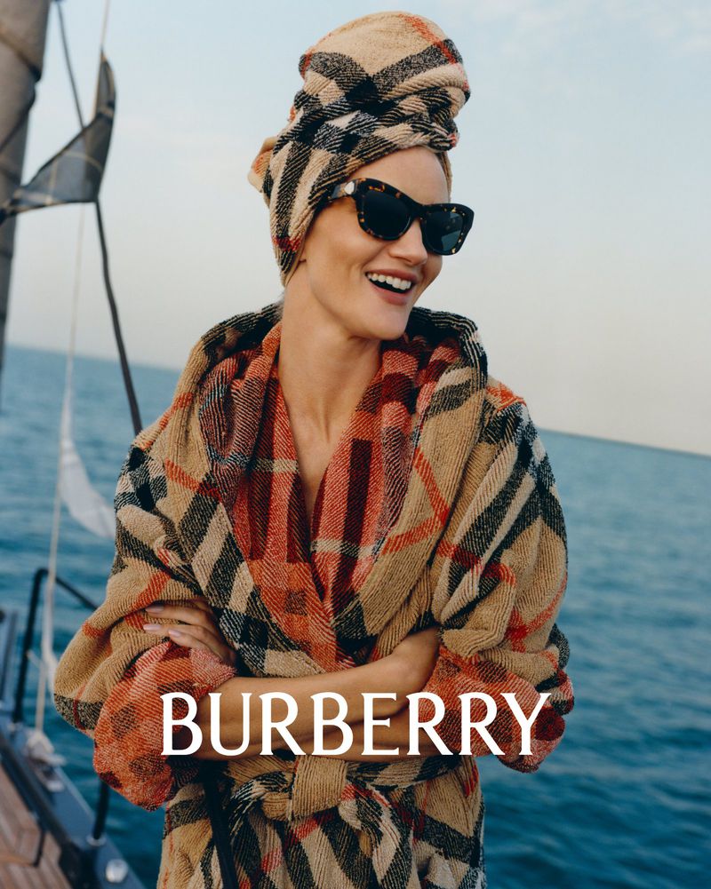 Smiling woman wearing a Burberry robe and head wrap stands on a boat with ocean and sky in the background; sunglasses, fashion.