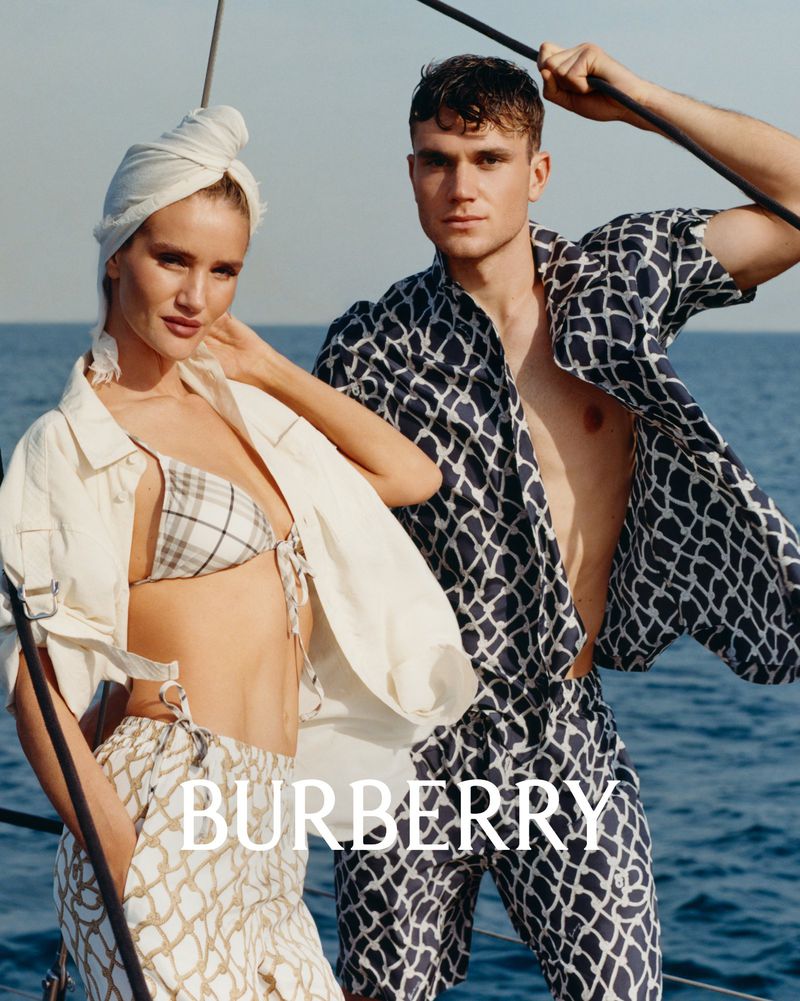 Two fashion models wear Burberry clothing on a boat, set against a tranquil ocean backdrop, with the Burberry logo visible.