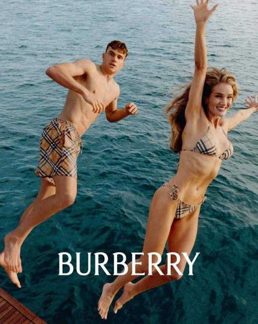 A man and woman jumping into blue water, both wearing swimwear with a distinctive plaid pattern. The text “BURBERRY” is at the bottom.