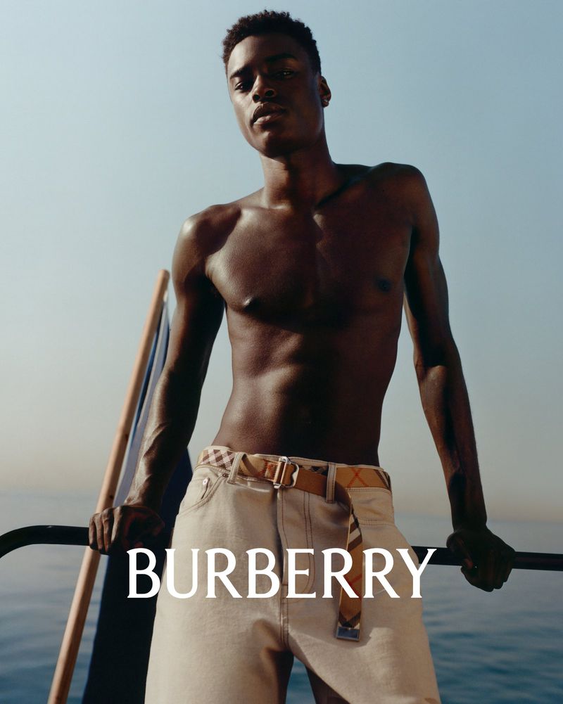 Fashion shot: A male model in light-wash jeans and a belt poses against a blue sky and ocean backdrop for a Burberry advertisement.