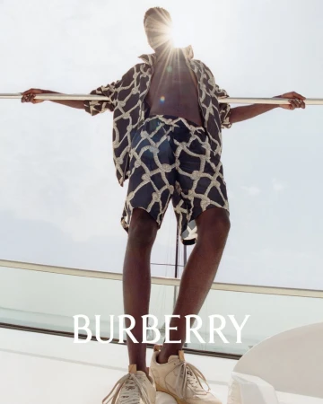A Black male fashion model stands on a yacht, wearing a patterned shirt and shorts, with a sunlit sky background and Burberry logo.