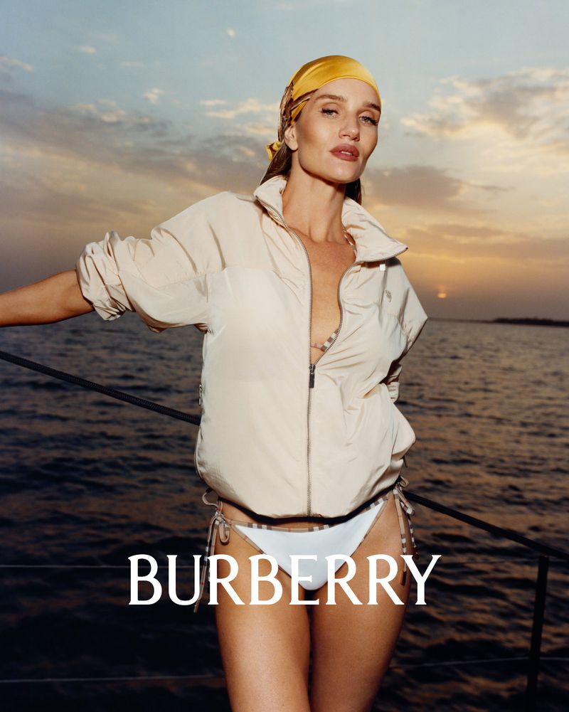 Model wearing a zip-up cream jacket and yellow headscarf standing on a boat deck at sunset