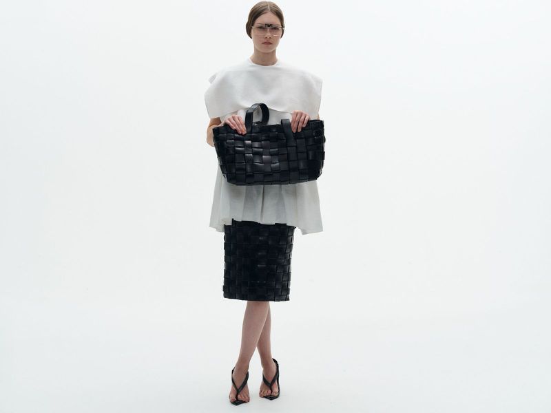 An adult female fashion model in a white top and woven black skirt stands in a white photo studio, holding a woven black tote bag.
