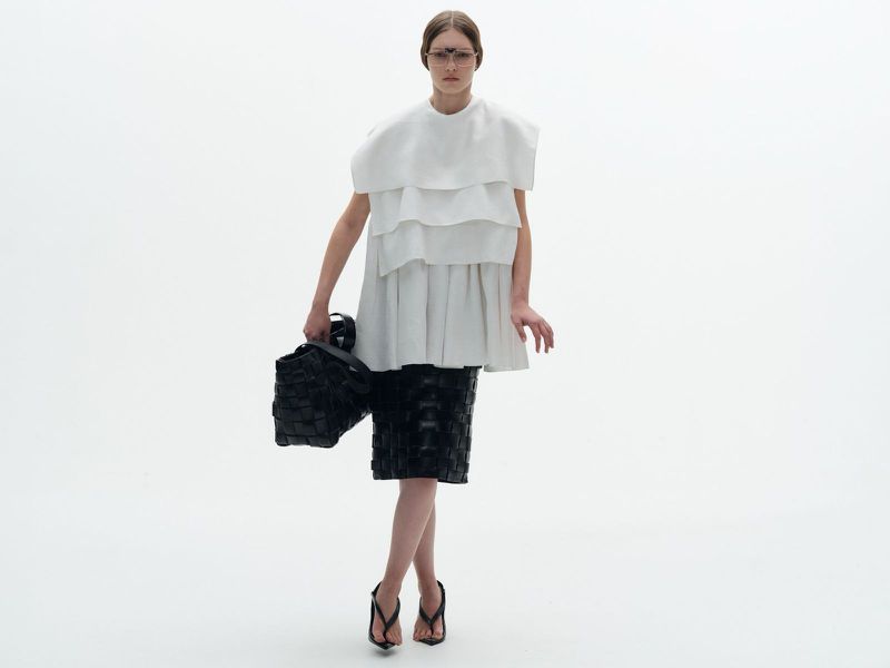 A woman in a white layered top and black skirt poses in a studio with a woven bag, creating a high-fashion and minimalist aesthetic.