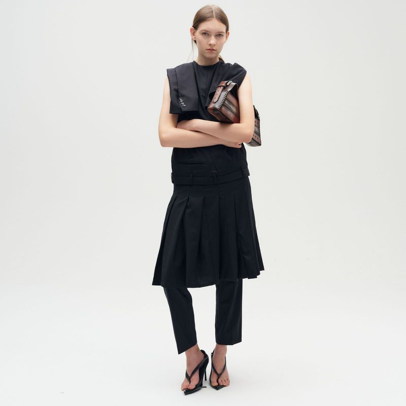 Woman posing in a dark, stylish sleeveless dress, long trousers, and heeled sandals against a bright backdrop, holding a striped purse.
