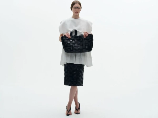 Model wearing a white structured top and woven black skirt holding a large black woven tote bag against a white seamless background