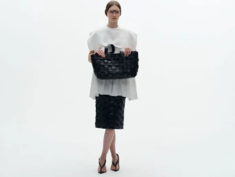 Model wearing a white structured top and woven black skirt holding a large black woven tote bag against a white seamless background