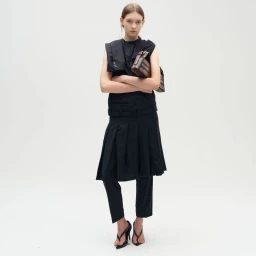 Model wearing a black sleeveless outfit with pleated skirt overlay and holding a structured handbag against a white background.