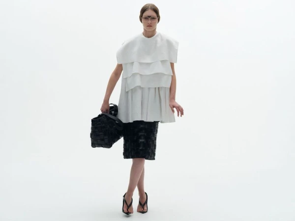 Model wearing a layered white top and black woven skirt holding a black handbag against a white background