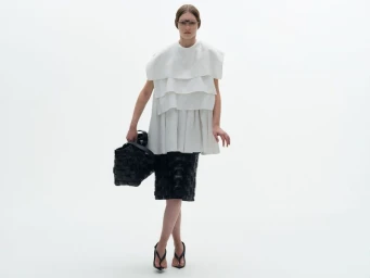 Model wearing a layered white top and black woven skirt holding a black handbag against a white background