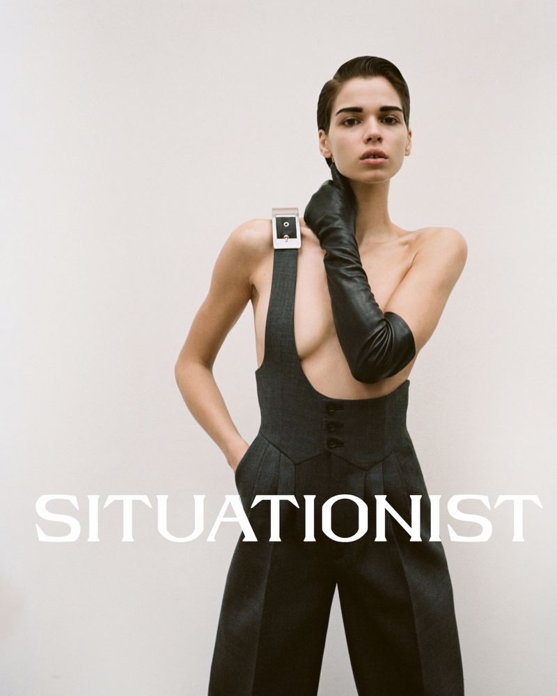 Model wearing a dark charcoal wool suspender garment and long black leather gloves against a white background.
