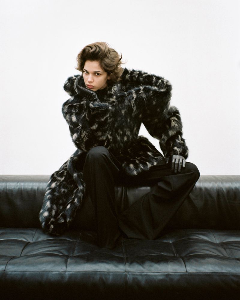 A woman sits on a black leather couch wearing a black and grey patterned fur coat and black pants in a studio setting.