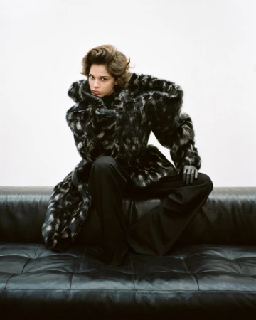 A woman sits on a black leather couch wearing a black and grey patterned fur coat and black pants in a studio setting.