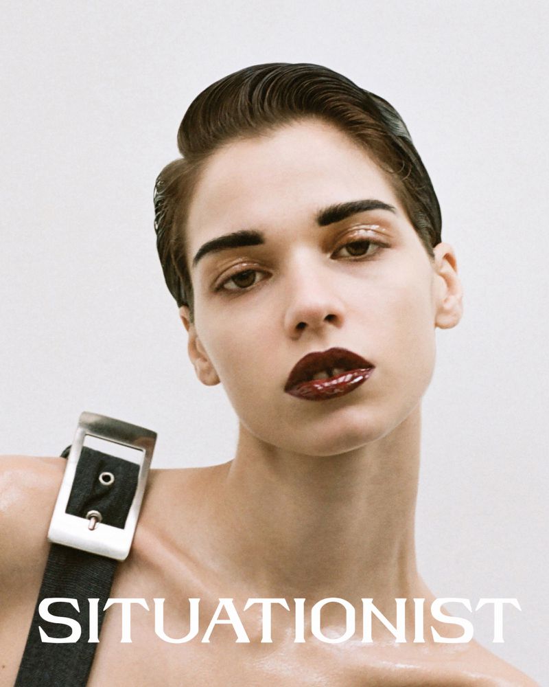 Close-up studio portrait of a woman with slicked-back hair and glossy dark red lips, wearing a wide belt strap, posing against a white background.