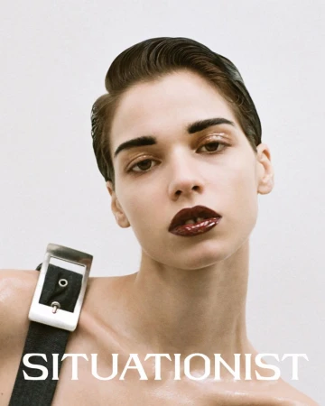 Close-up studio portrait of a woman with slicked-back hair and glossy dark red lips, wearing a wide belt strap, posing against a white background.