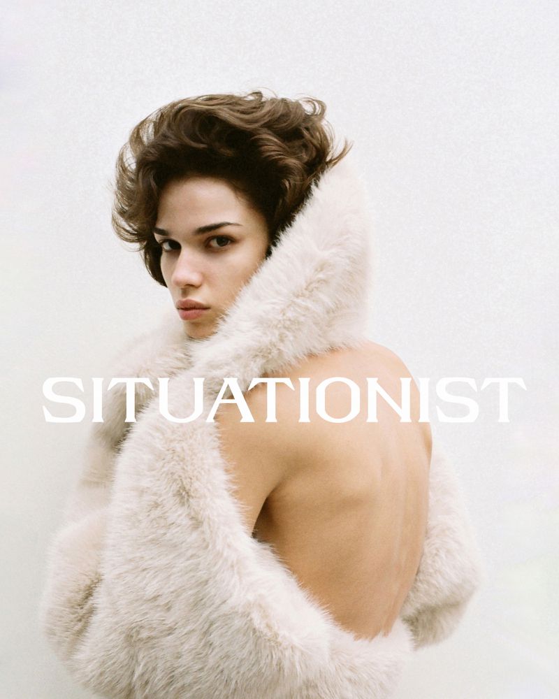 Fashion portrait of a person with brown hair posing indoors, wearing a fur-lined coat against a white wall with 'SITUATIONIST' text.