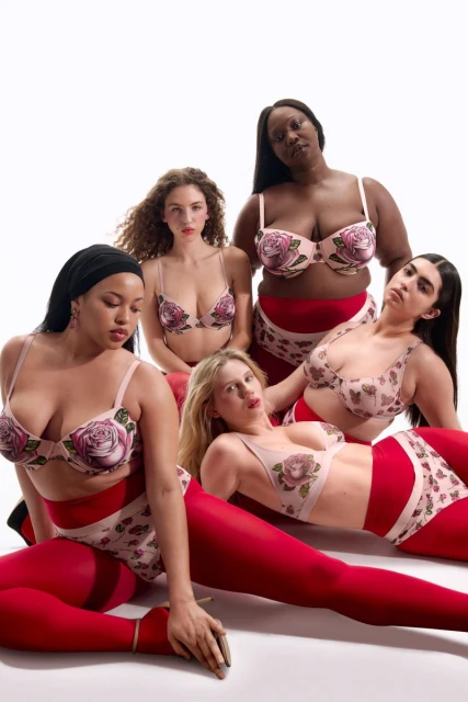Five women pose in a photo studio, wearing matching floral print lingerie and red tights, set against a bright white backdrop.
