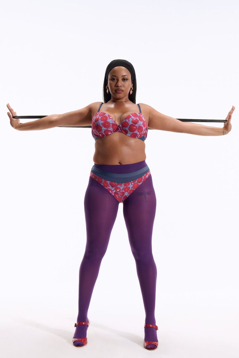 Adult woman in a bra and tights standing in front of a white background holding an exercise band out with both arms.