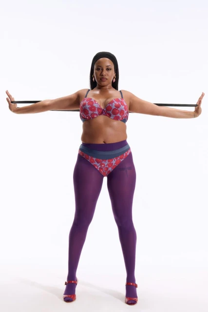 Adult woman in a bra and tights standing in front of a white background holding an exercise band out with both arms.