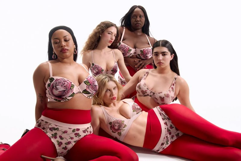 Five diverse women pose in matching floral lingerie and red tights against a white backdrop in a studio setting.