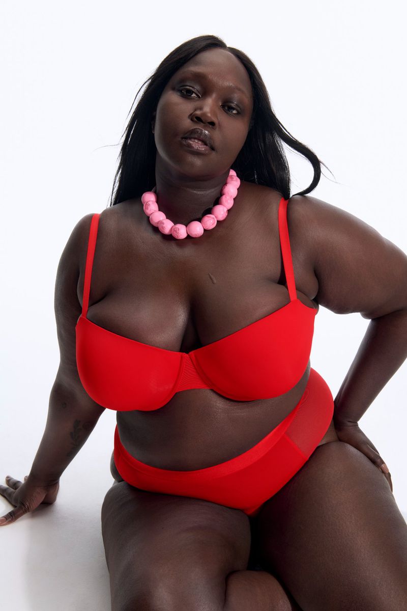 Adult female poses in a bright red bra and matching underwear set, accessorized with a pink beaded necklace against a white backdrop.