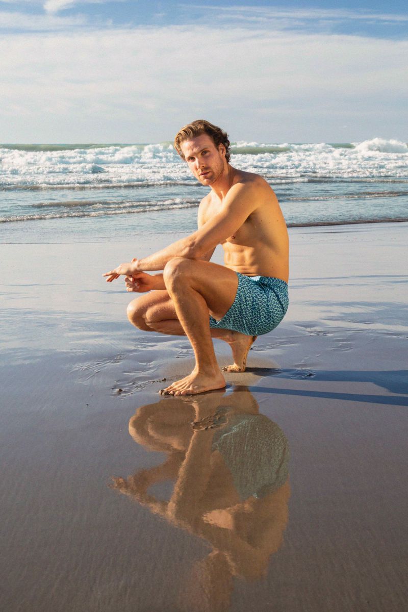 Man in swim trunks squats on wet sandy beach with ocean waves and blue sky. His reflection shows on wet sand.