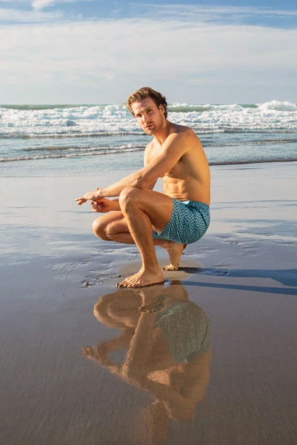 Man in swim trunks squats on wet sandy beach with ocean waves and blue sky. His reflection shows on wet sand.
