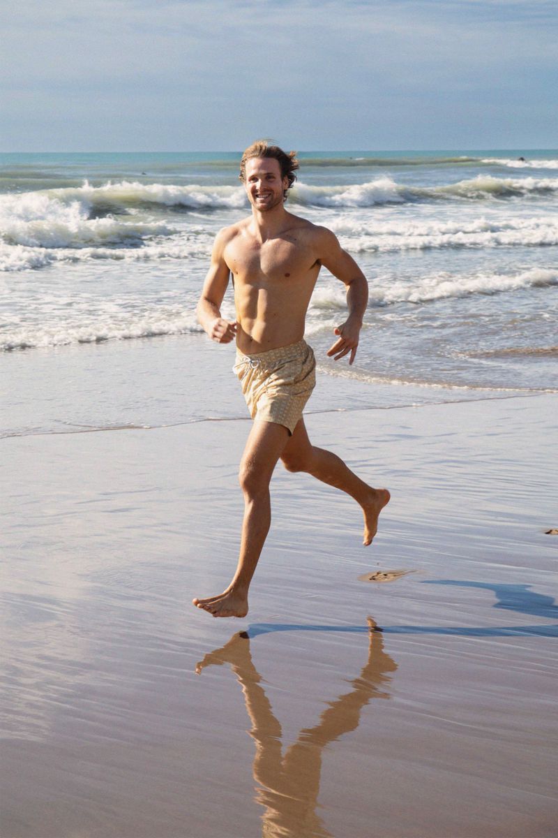 Athletic man running barefoot on a wet sandy beach along the ocean shoreline under a partly cloudy sky on a sunny day.
