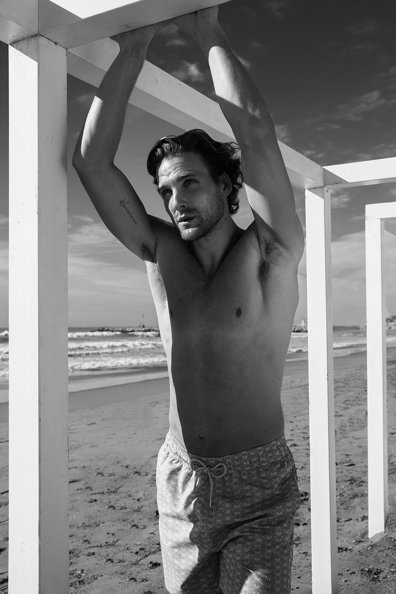 Black and white portrait of a shirtless man in patterned swim trunks, standing on a beach and leaning on a white structure.