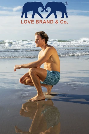 Male model posing on a sandy beach near the ocean wearing swimwear for Love Brand & Co. Fashion shot with ocean background.