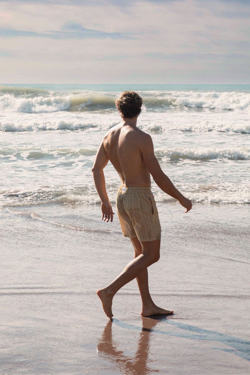 Bare-chested man in patterned swim trunks walks barefoot along a sandy beach with waves and cloudy skies in the background.