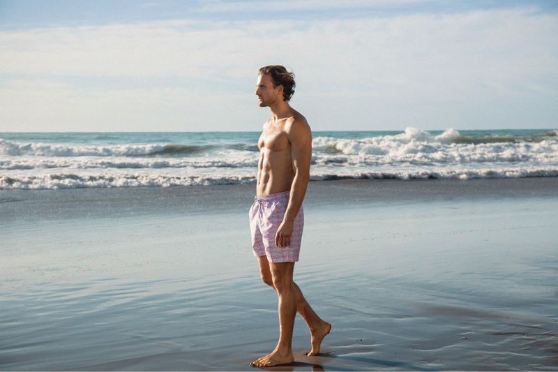 Athletic man walking barefoot on a wet sandy beach with waves and blue sky in the background during daytime, wearing patterned swim trunks.