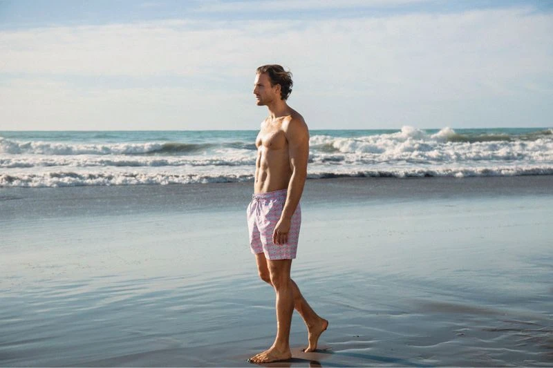 Athletic man walking barefoot on a wet sandy beach with waves and blue sky in the background during daytime, wearing patterned swim trunks.